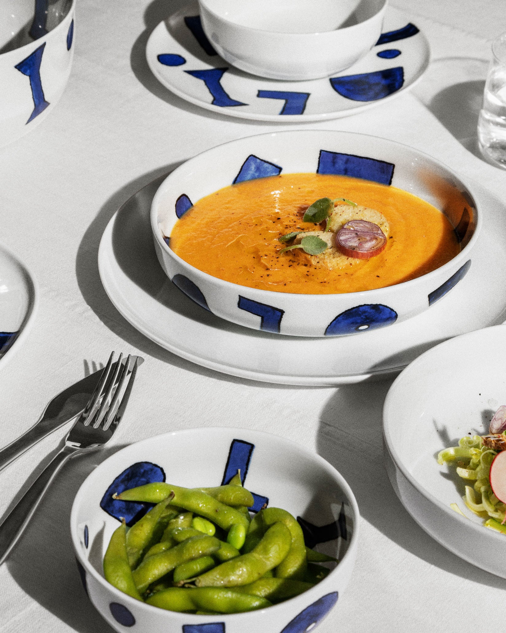 White ceramic table sets by Alessi with blue geometric patterns, holding soup and edamame on a white tablecloth.