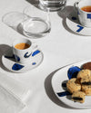 White ceramic table set by Alessi with hand-painted blue abstract patterns on a white tablecloth.