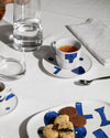 White ceramic table set with blue abstract patterns by Alessi, featuring cups, saucers, and a platter with drinks and cookies.
