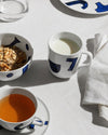 White ceramic table set by Alessi with bold blue abstract patterns, serving breakfast on a linen tablecloth.