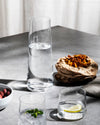 Tall clear glass pitcher by Alessi filled with water, styled on a grey table with smaller glasses and snacks.