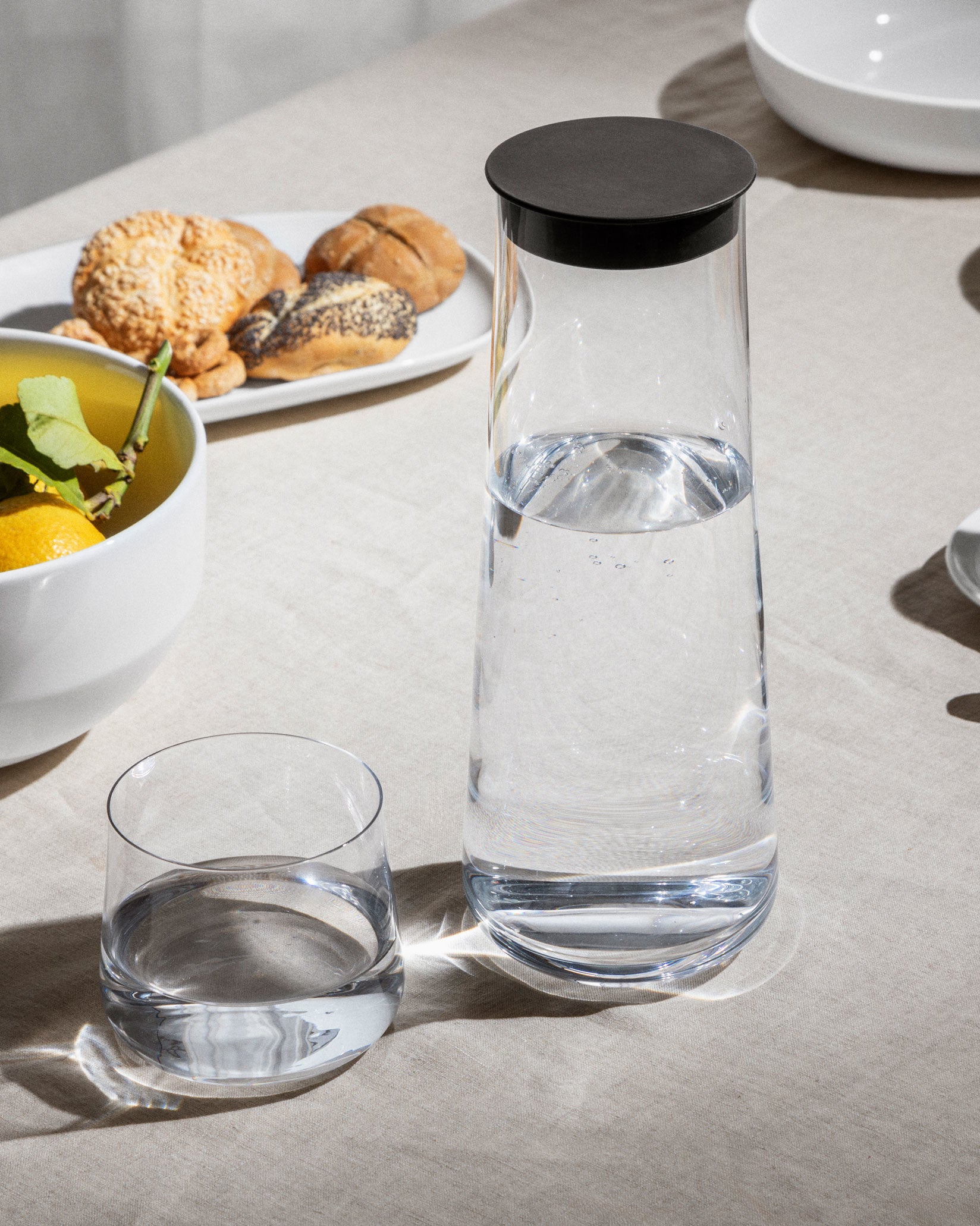 Clear glass pitcher with a black lid by Alessi, filled with water, alongside a drinking glass and bread rolls.