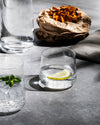Clear glass water glasses by Alessi, with one holding a lemon slice and another mint, on a grey surface with snacks.