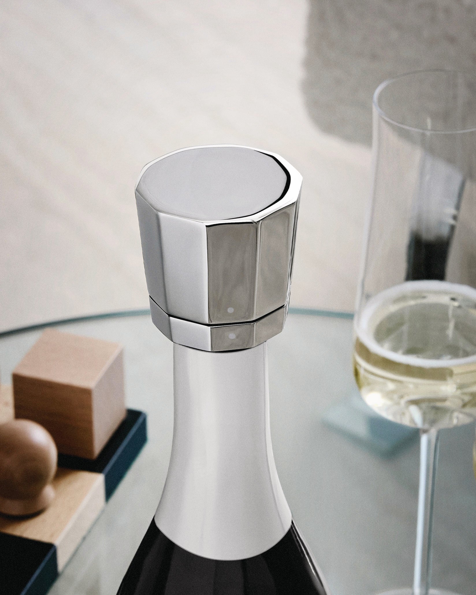 Silver faceted stopper by Alessi on a white champagne bottle next to a glass.