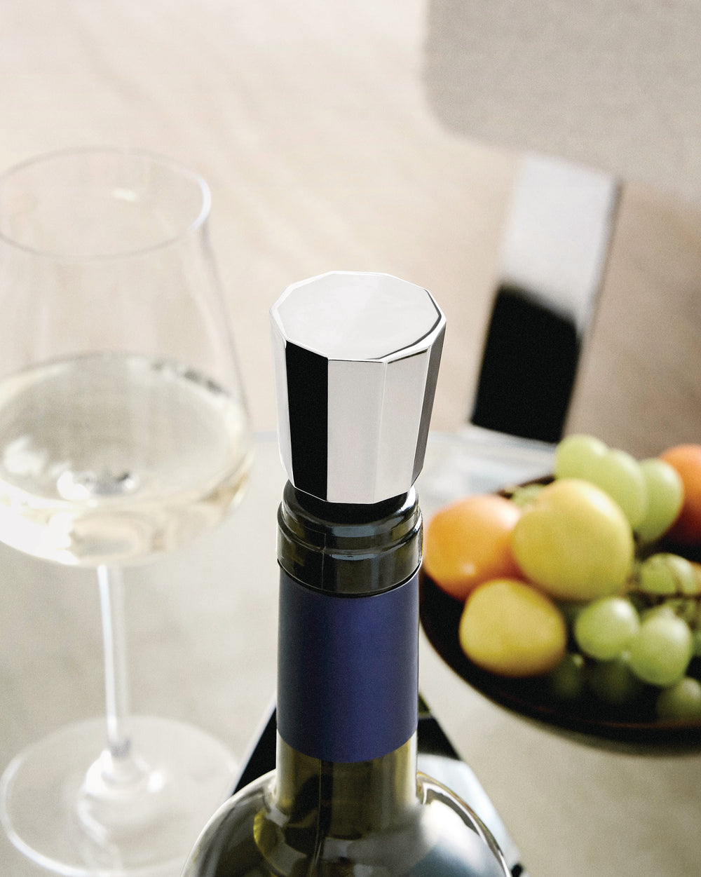 Silver hexagonal stopper by Alessi on a wine bottle, beside a glass of white wine and fruit.