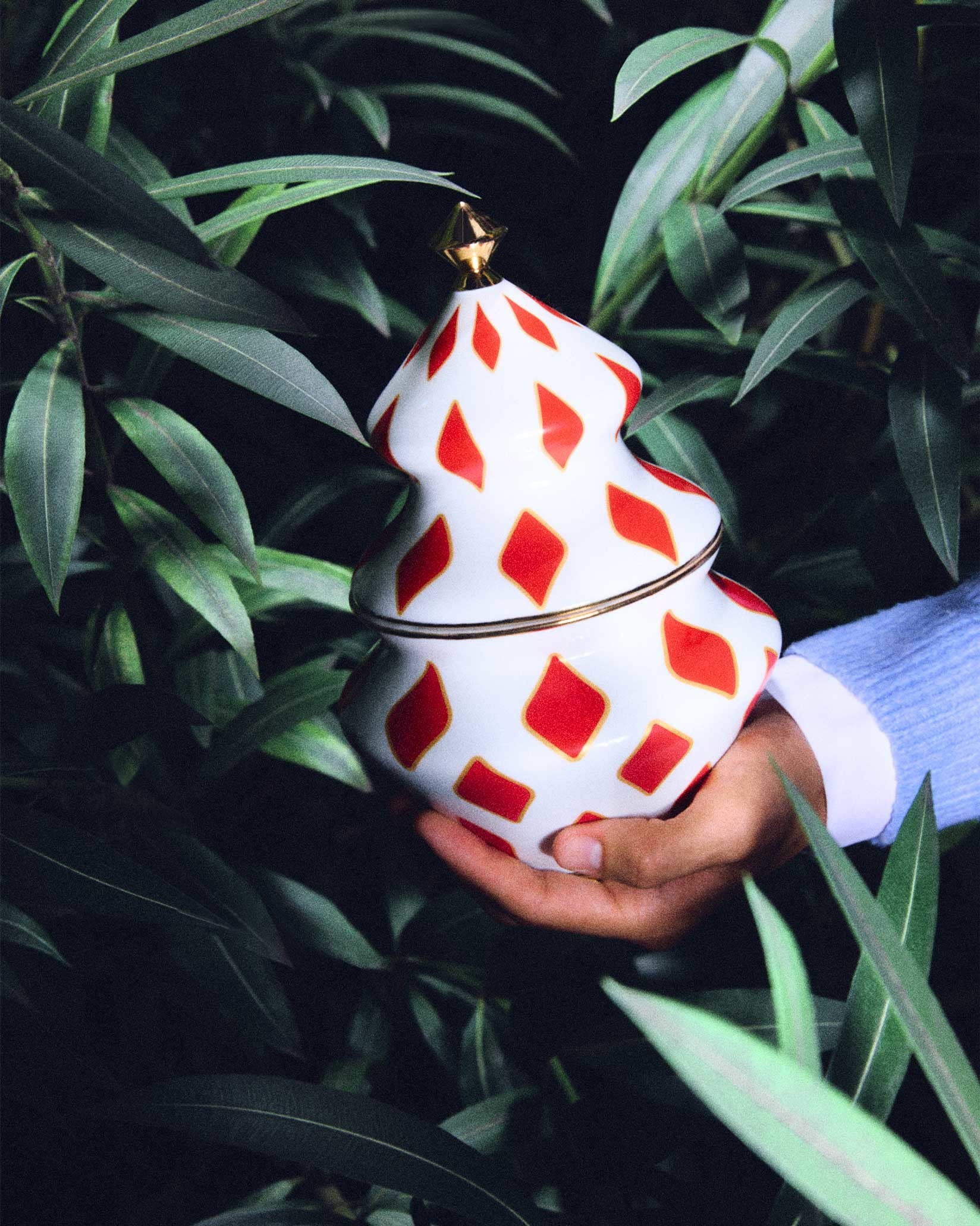 White and red ceramic Christmas tree decoration by Alessi, featuring a diamond pattern and a gold top, held in a hand amidst green leaves.