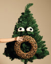 Playful googly eye and wicker wreath Christmas tree ornaments by Alessi, on a green tree, held by a hand.