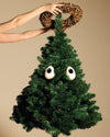Brown twig wreath by Alessi being placed by hands atop a green artificial Christmas tree with googly eyes.