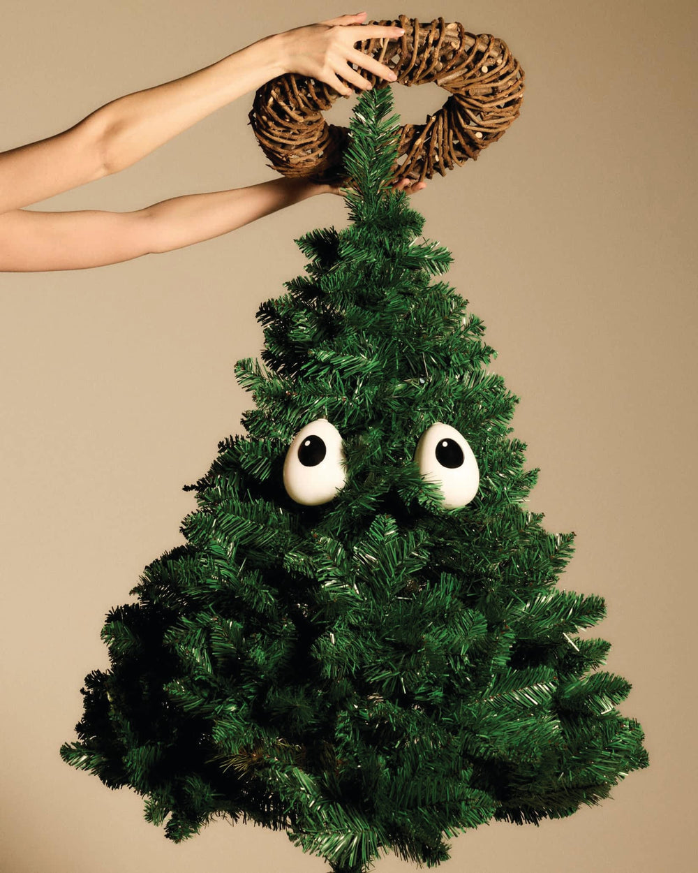 Brown twig wreath by Alessi being placed by hands atop a green artificial Christmas tree with googly eyes.
