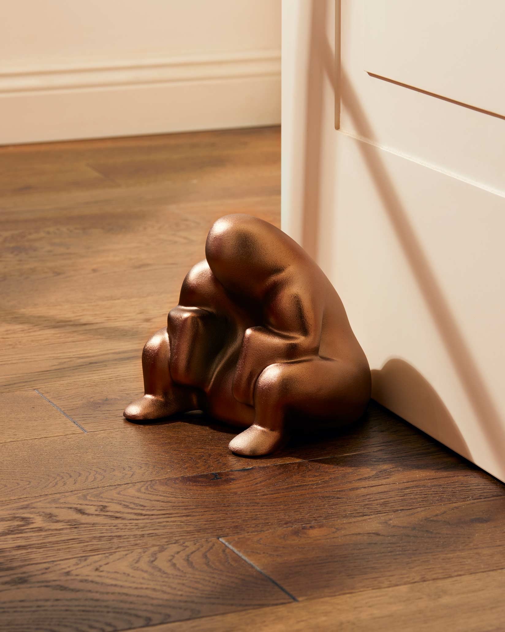 Bronze-colored doorstop by Alessi, shaped like a stylized crouching figure on a wooden floor propping a white door.