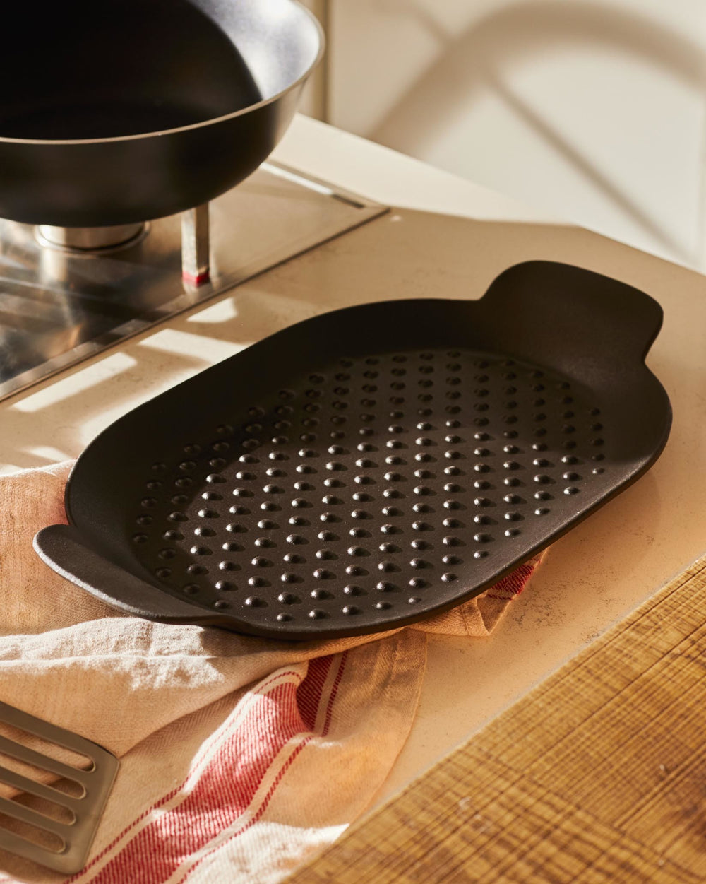 Black oval grill pan by Alessi with a dimpled surface, resting on a striped kitchen towel.