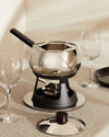 Mami fondue and bourguignonne set by Alessi, with a polished stainless steel pot, black cast iron stand, and lid, presented on a linen tablecloth.