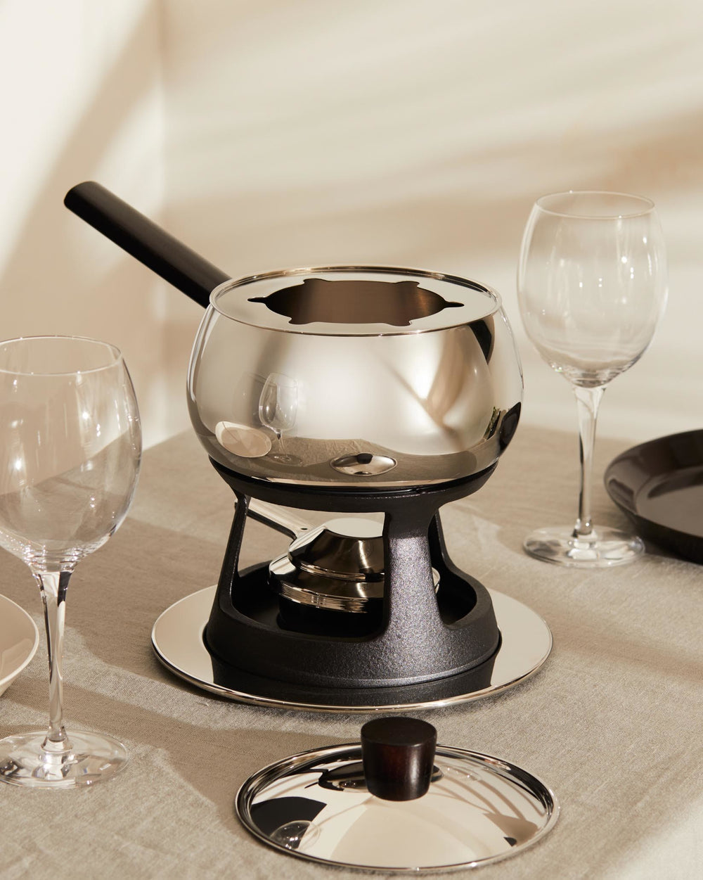 Mami fondue and bourguignonne set by Alessi, with a polished stainless steel pot, black cast iron stand, and lid, presented on a linen tablecloth.