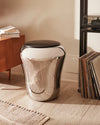 Chrome laundry basket by Alessi with a black top, featuring a sleek, reflective, and curved design on a wood floor.