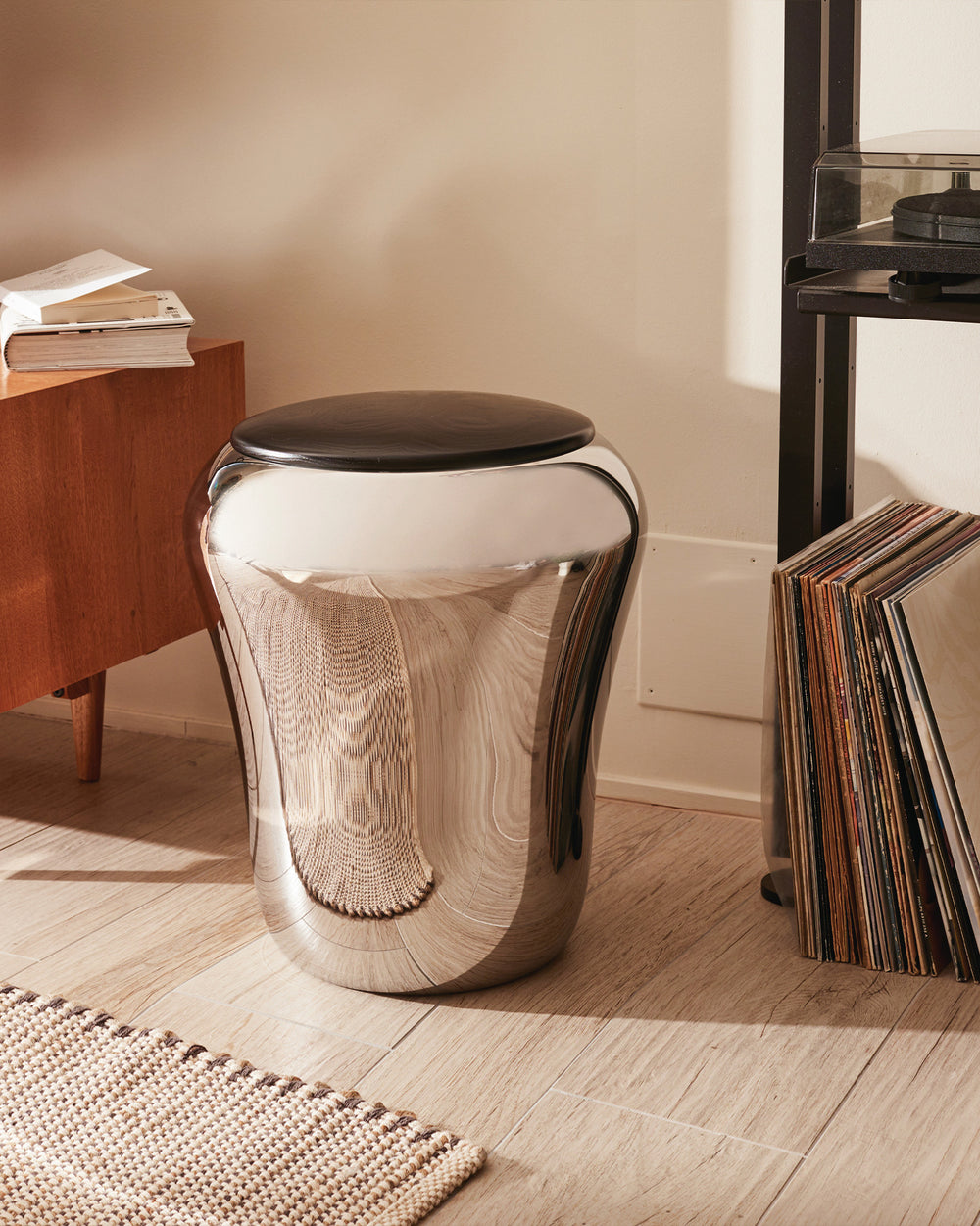 Chrome laundry basket by Alessi with a black top, featuring a sleek, reflective, and curved design on a wood floor.