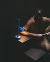 Blue table lamp by Alessi with a minimalist design, casting a warm glow on a man and a gold surface.