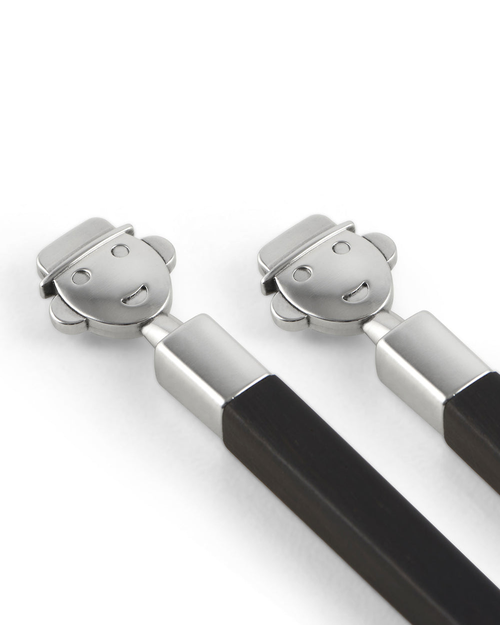 Pair of brushed metal and dark wood sushi sticks by Alessi, with smiling figure toppers, angled on a white surface.