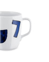 White ceramic mug by Alessi with a hand-painted blue number 7 and a partial blue design, cropped side view.