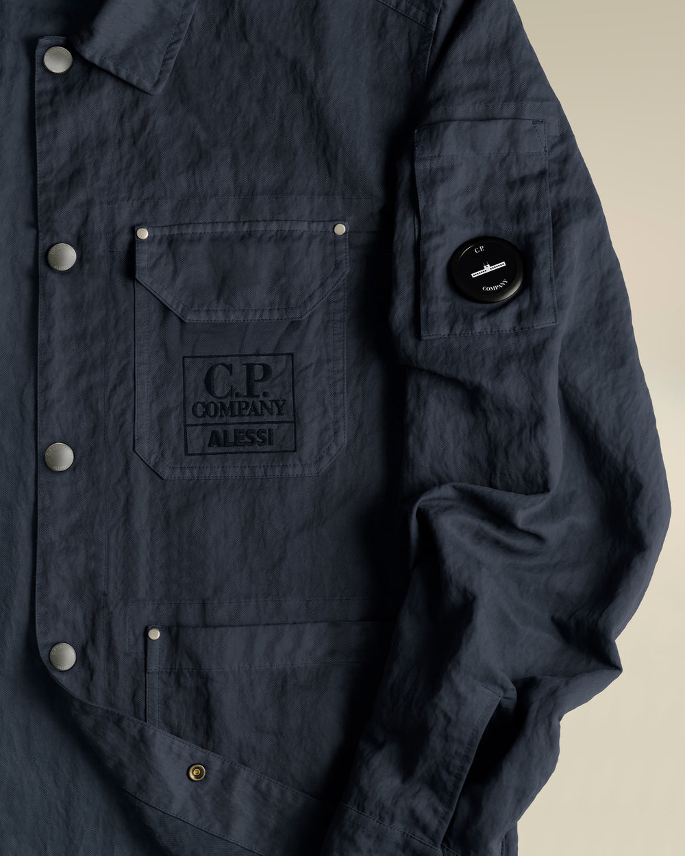 Dark blue jacket by Alessi with C.P. Company branding, snap buttons, and a sleeve lens detail.