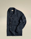 Dark blue crinkled utility jacket by Alessi, with multiple pockets, C.P. Company logos, and a sleeve lens badge, laid flat.