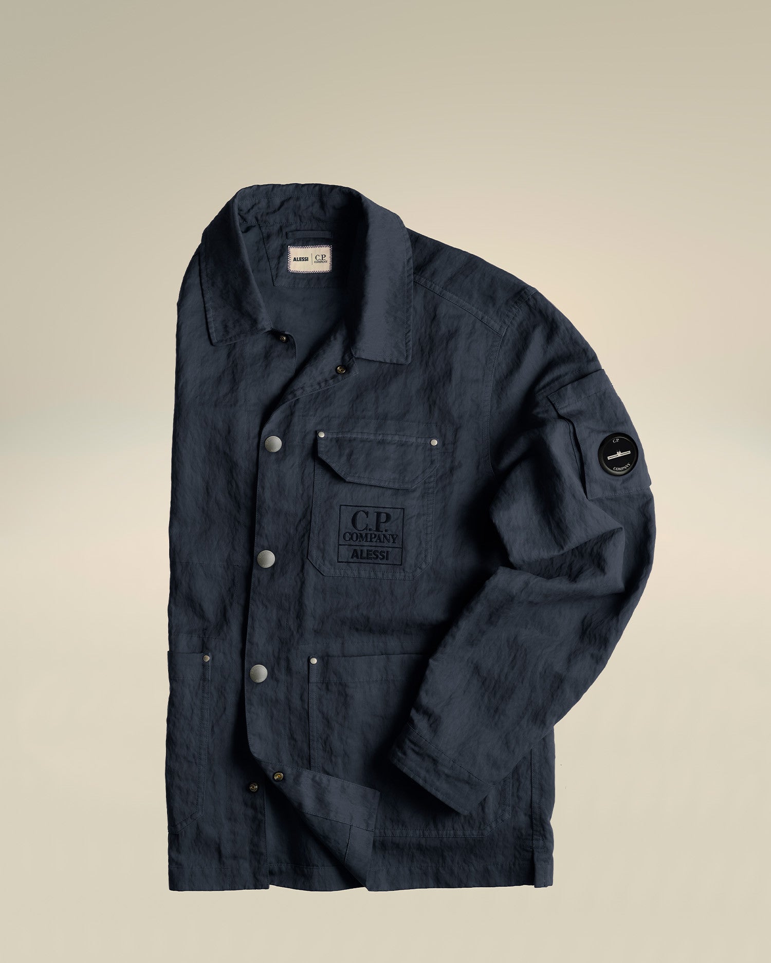 Dark blue crinkled utility jacket by Alessi, with multiple pockets, C.P. Company logos, and a sleeve lens badge, laid flat.