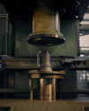 A matte silver metal espresso maker by Alessi, positioned under a large industrial press.