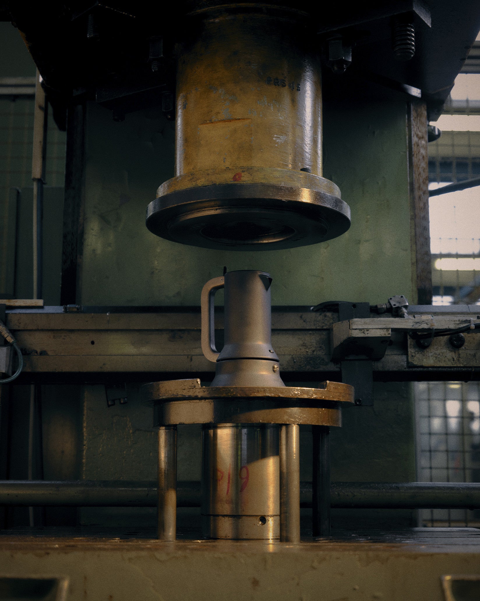 A matte silver metal espresso maker by Alessi, positioned under a large industrial press.