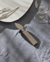 Silver oyster knife by Alessi with a fish scale patterned handle, resting on a blue linen tablecloth.