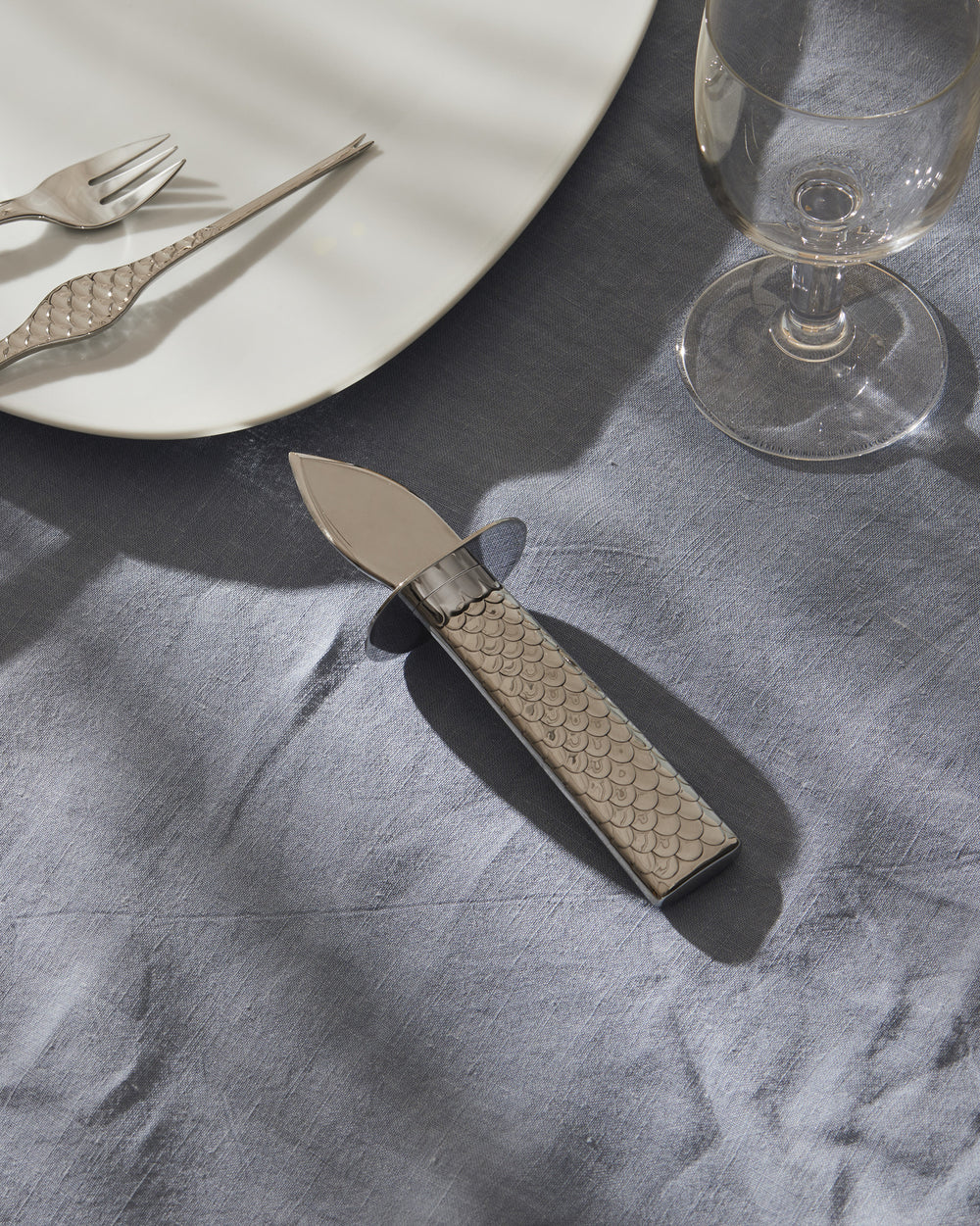 Silver oyster knife by Alessi with a fish scale patterned handle, resting on a blue linen tablecloth.