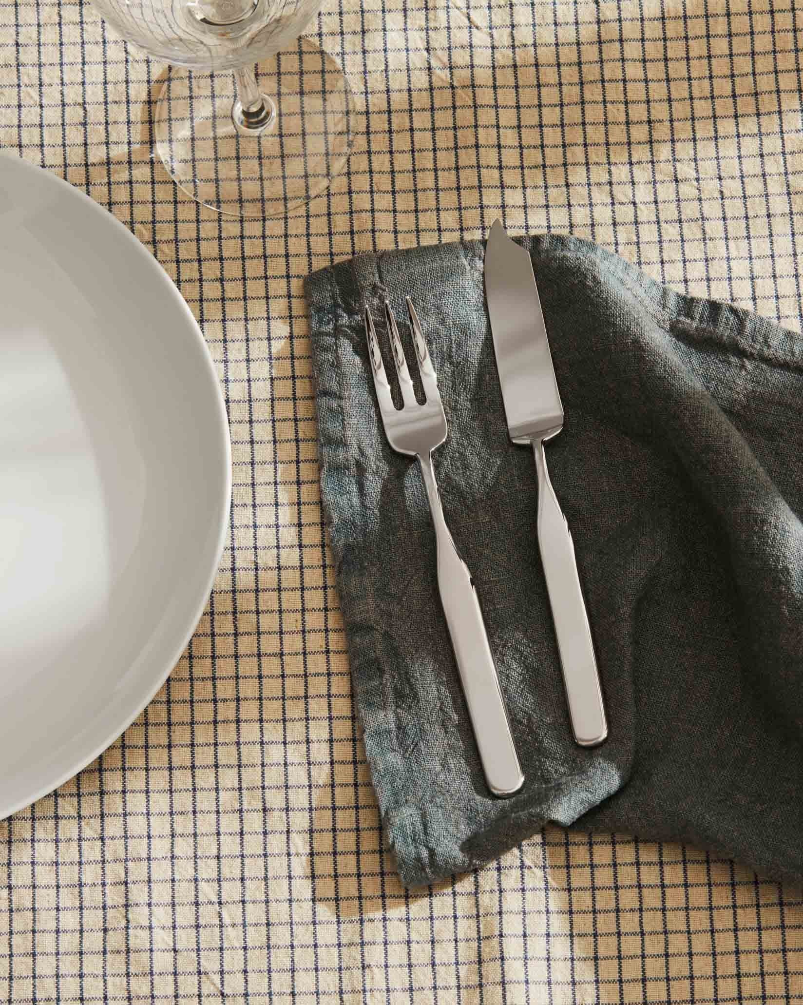 Silver fish fork by Alessi, with a matching knife on a grey linen napkin, atop a plaid tablecloth.