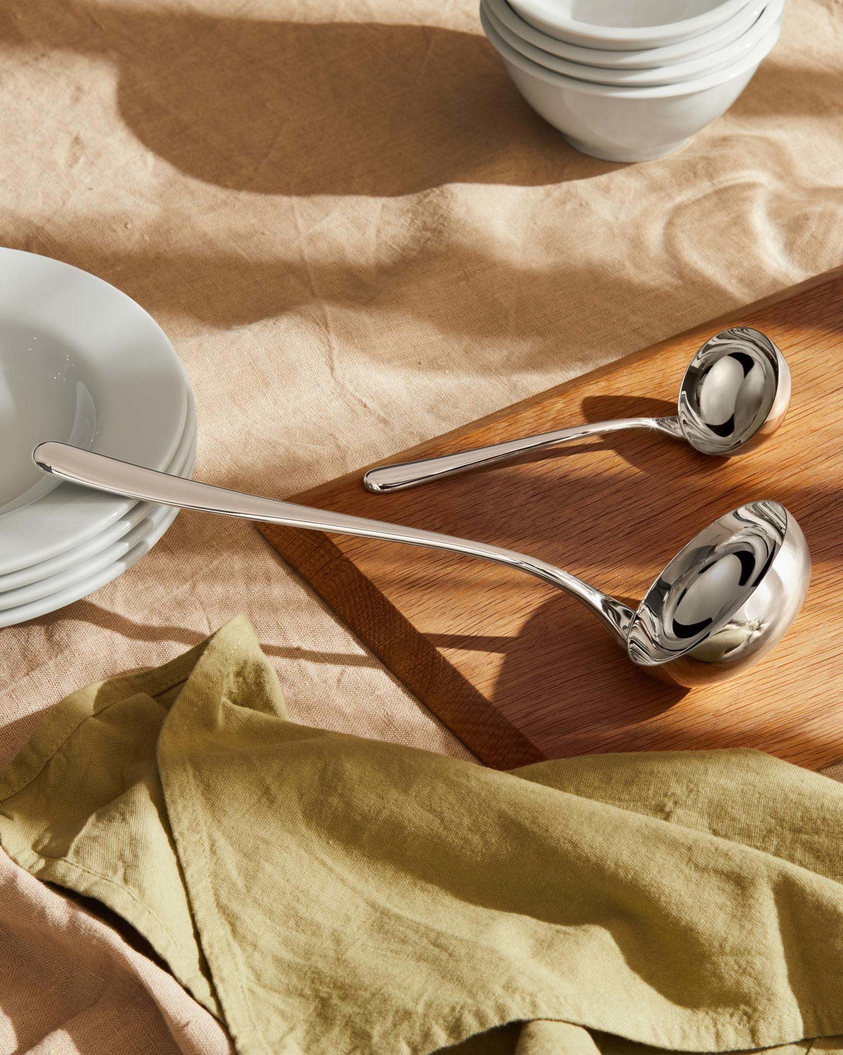 Two polished silver-toned ladles by Alessi, resting on a wooden board with white dishes and a green napkin.
