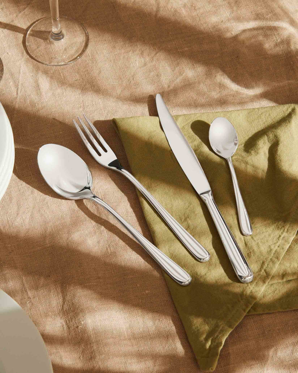Polished silver flatware set by Alessi, with striped handles, including a knife, fork, and two spoons, on green and brown linen.