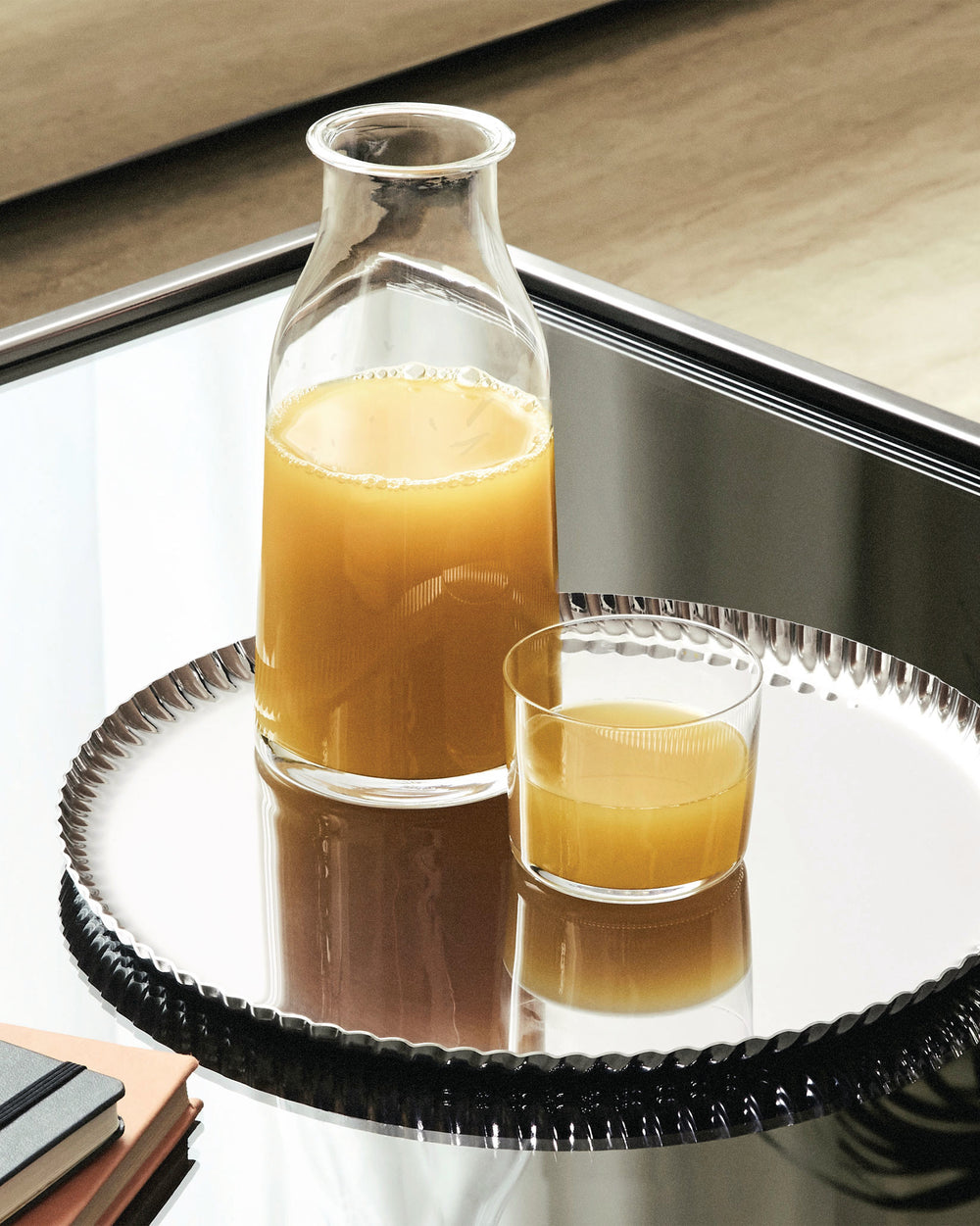 Silver oval tray by Alessi with a ruffled edge, holding a bottle and glass of orange juice on a mirrored table.