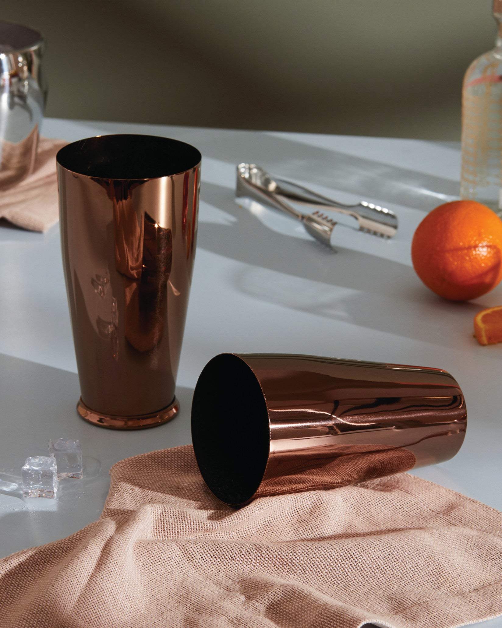 Copper cocktail shakers by Alessi, one upright and one lying on a grey counter with ice and an orange.