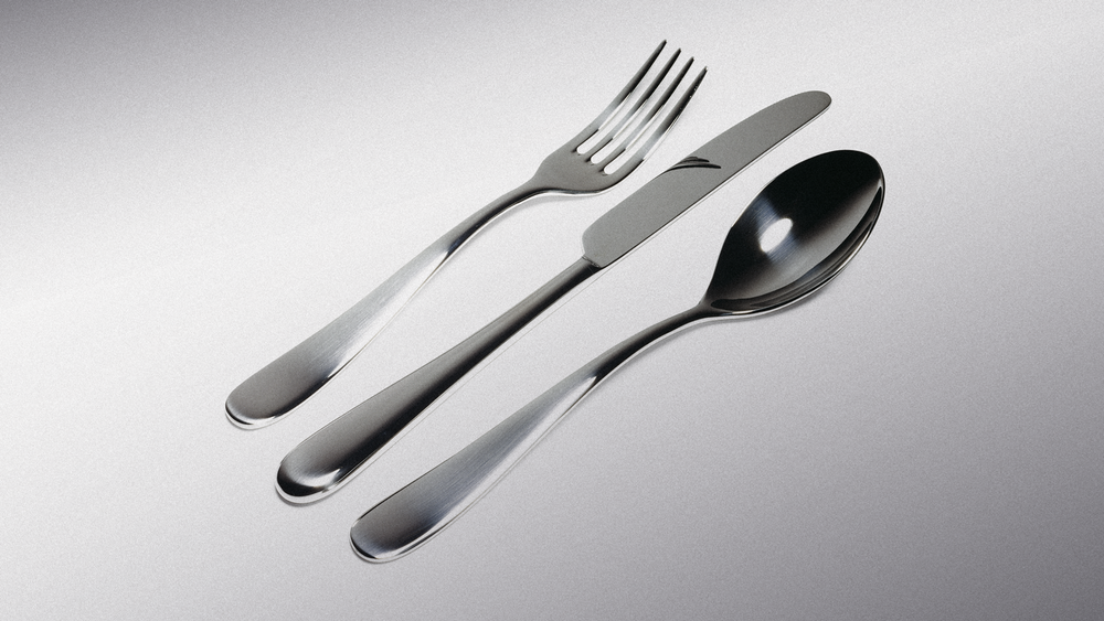 Shiny silver fork, knife, and spoon arranged diagonally on light gray surface, reflective surfaces, minimal lighting.