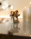 White ornamental figurines "Do, Re, Mi" by Alessi playing musical instruments, on a reflective surface with lit candles.
