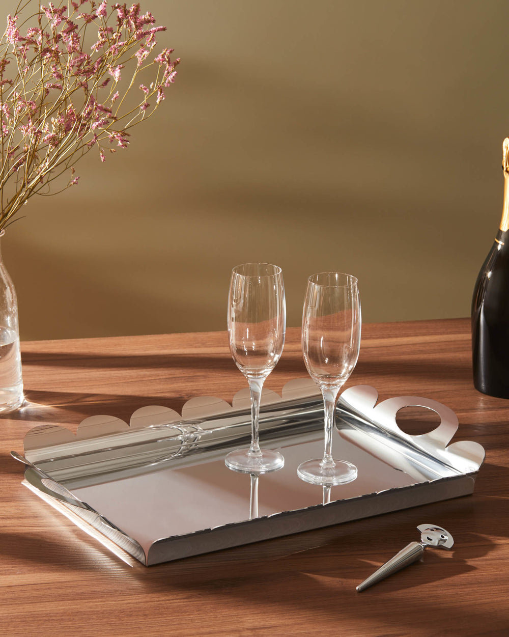A rectangular silver tray from Alessi with a scalloped edge and raised sides, holding two champagne flutes.