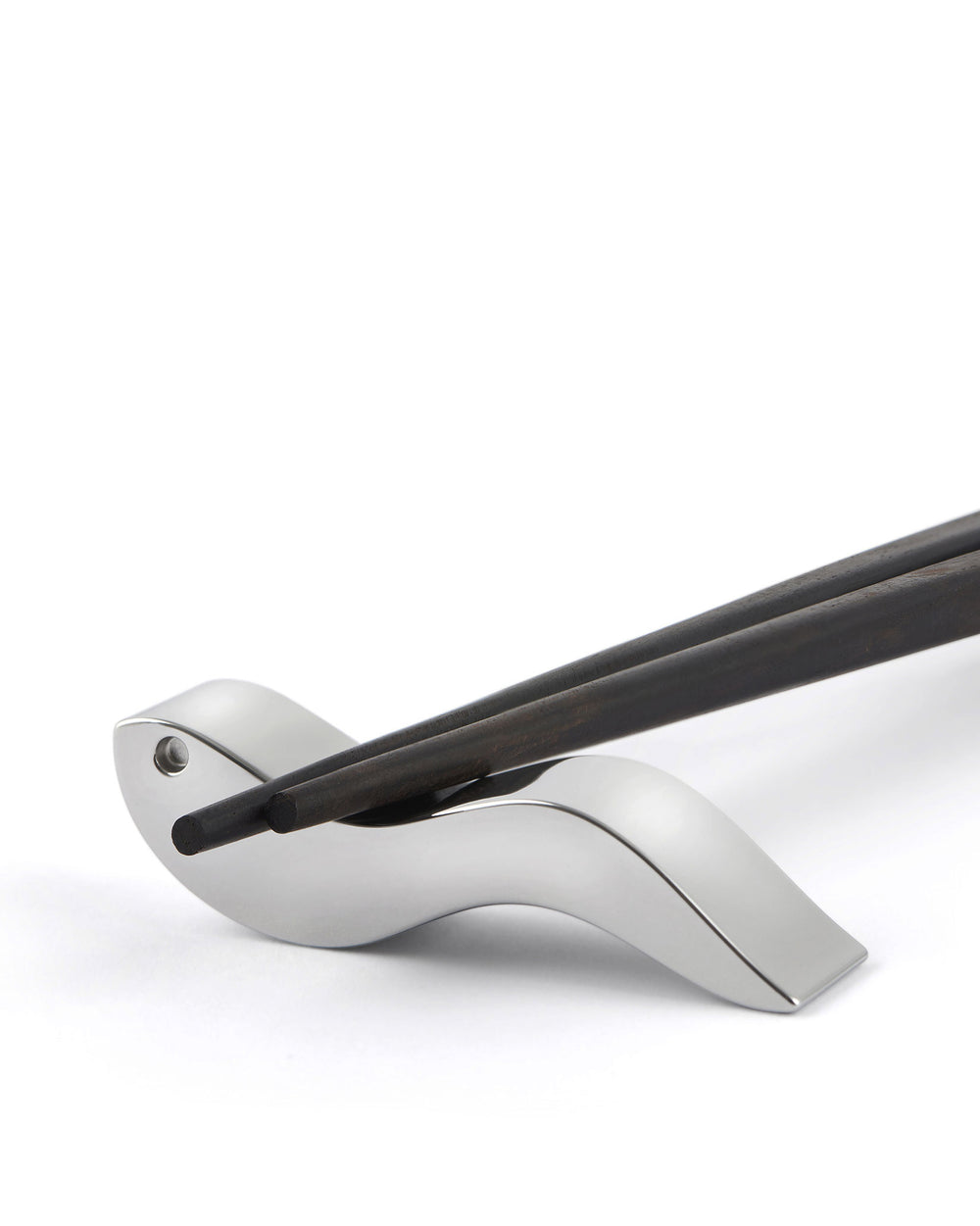 Silver metallic chopstick rest by Alessi holding two dark wooden chopsticks on a white background.