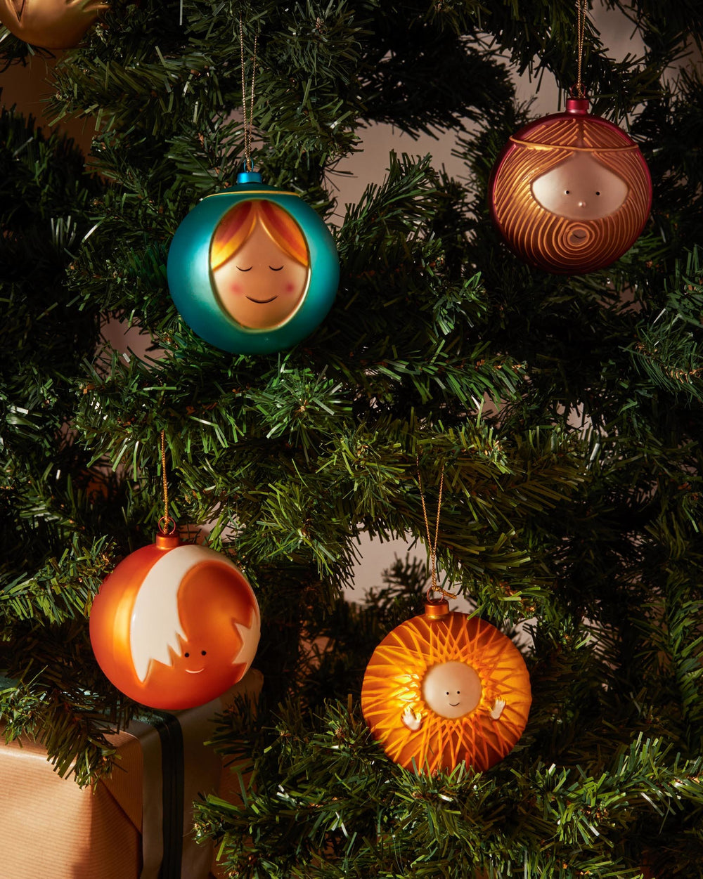 Glowing orange Christmas tree ornament by Alessi, with a central smiling face, small hands, and radiating lines, hanging on a green tree.
