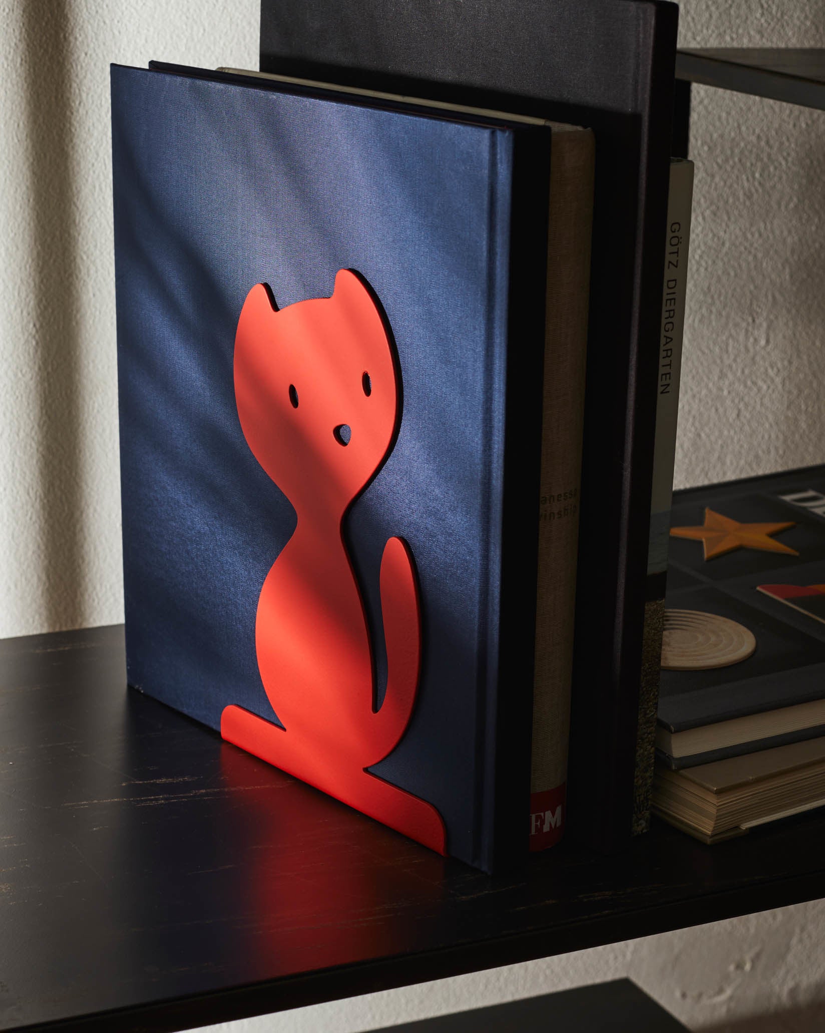 Red cat-shaped bookstand by Alessi, holding a dark blue textured book on a black shelf with striped shadow pattern.
