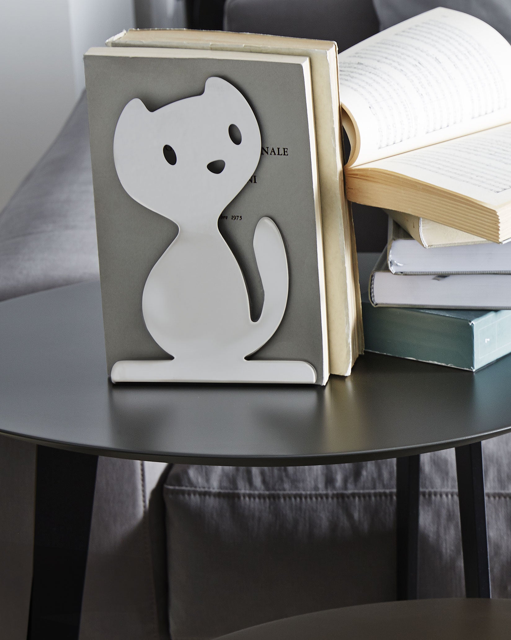 White cat-shaped bookend by Alessi, holding open books on a dark gray table in a living room.