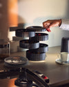 Dark grey fluted capsule holder by Alessi, stacked with multiple tiers, as a hand retrieves a red capsule.