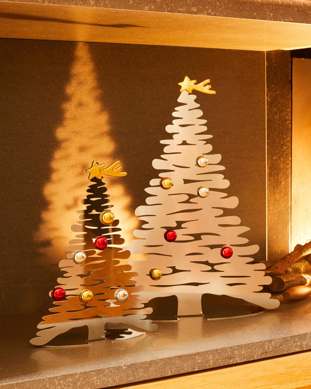 Two gold metallic Christmas tree ornaments by Alessi, with abstract cutout branches, shooting stars, and colorful baubles.