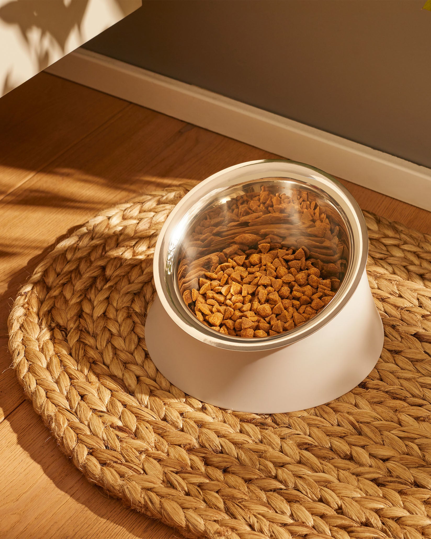 Elevated angled dog bowl by Alessi, with a stainless steel inner and white stand, filled with brown kibble, on a braided straw mat.