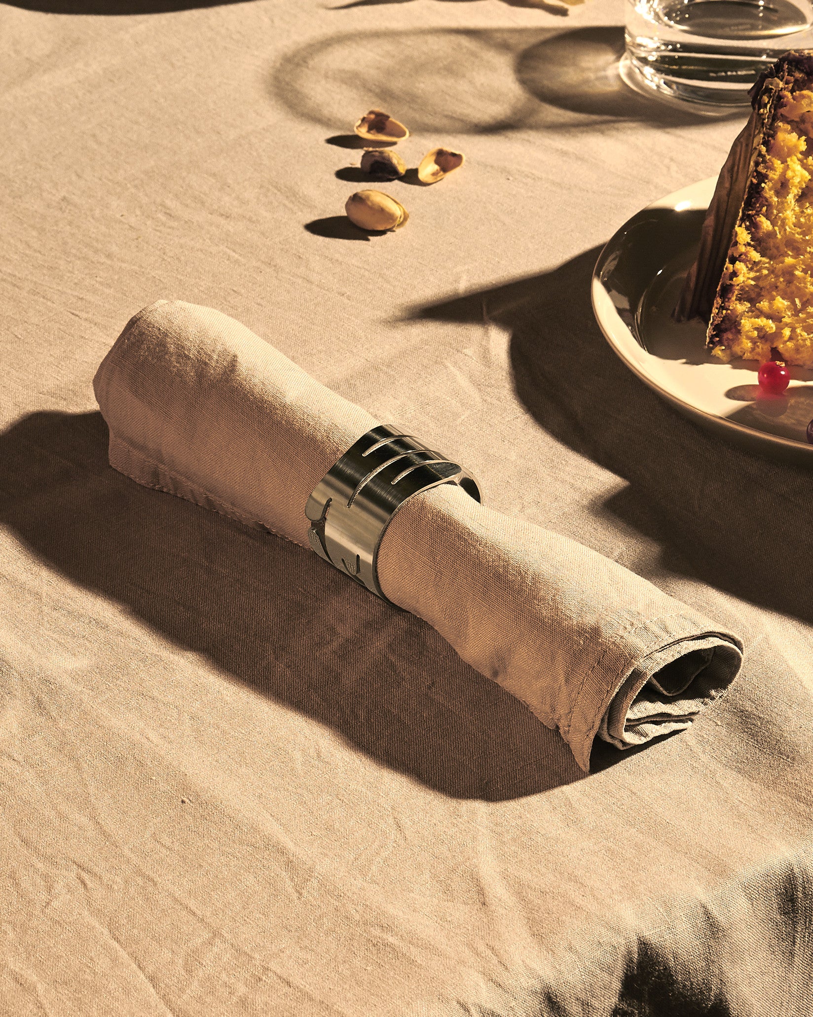 Brushed silver metal napkin holder by Alessi featuring a laser-cut bark-like pattern, holding a beige linen napkin.