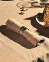 Silver-toned metal napkin holder by Alessi with a cut-out pattern, embracing a beige linen napkin on a table.