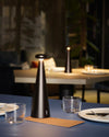 Dark bronze cone-shaped table lamp by Alessi on a blue table set with dinnerware, glowing warmly.