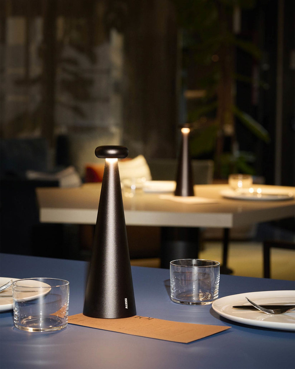 Dark bronze cone-shaped table lamp by Alessi on a blue table set with dinnerware, glowing warmly.