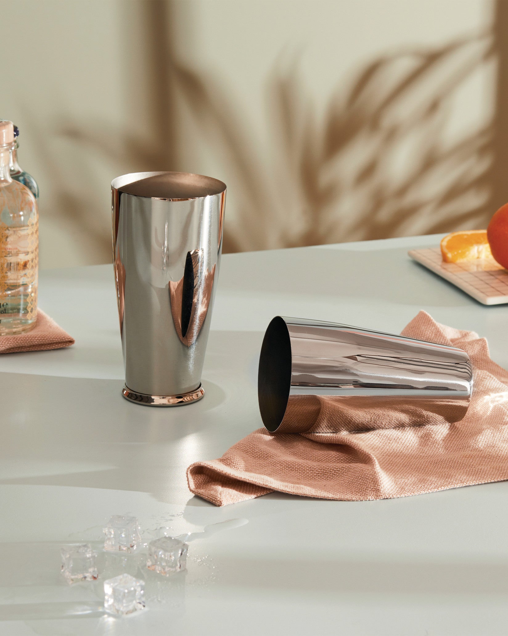 Polished stainless steel and copper cocktail shakers by Alessi, one upright and one lying on a peach textured cloth, with ice cubes.