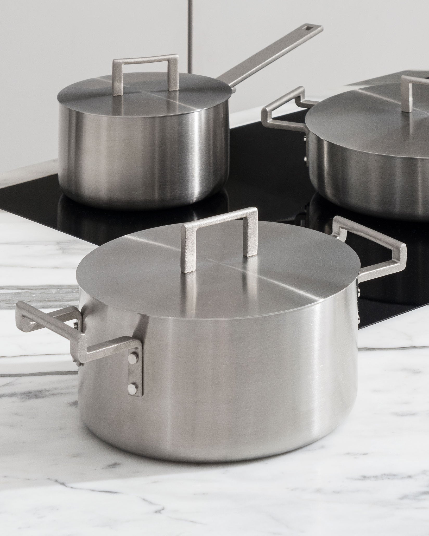 Brushed stainless steel pots by Alessi with minimalist square-loop handles and flat lids on a stove.